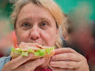 Retired elderly woman with red hair is tasting large hamburger cooked in high-tech kitchen. Master class on cooking meat dishes and hamburgers