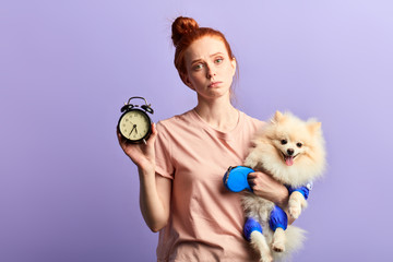 beautiful young sleepy unhappy woman holding a clock and her dog in hands, she doesn't want to walk...