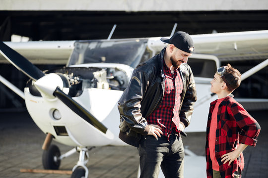 Little boy dressed in red shirt, jeans and aviator glasses looks with smile at his flight coach, full of enthusiasm, ready to start lesson on white single-engine propeller airplane. Outdoor shot.