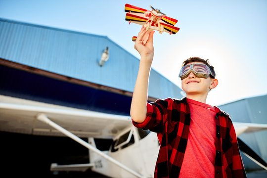 view of lovely kid playing with toy plane outdoor near the shed, white single-engine aircraft stands behind him, wears aviator glasses, red shirt and jeans. Aeromodelling hobby. Outdoor shot.