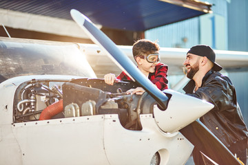 teenager boy who dreams to be an aircraft engineer and his dad pilot look at each other and laugh loudly, standing near white light propeller airplane, spending great time together.