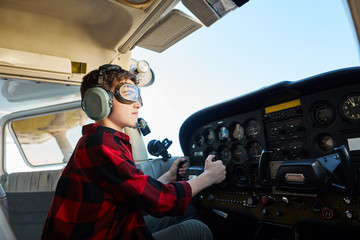 portrait of boy looking focused and serious, sitting in cockpit of small propeller airplane, wears...