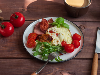 Rich breakfast of omelet with grilled sausage and fresh tomatoes and fresh atromatous herbs, cutlery on a wooden tray