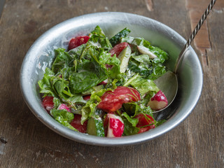 Fresh summer salad of tomatoes, cucumbers and aromatic herbs in a deep bowl with a spoon, with sour cream on an old wooden table