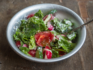 Fresh summer salad of tomatoes, cucumbers and aromatic herbs in a deep aluminum bowl with a spoon, with sour cream on an old wooden table