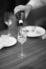 The waiter pours white sparkling wine in a tall glass standing on the table