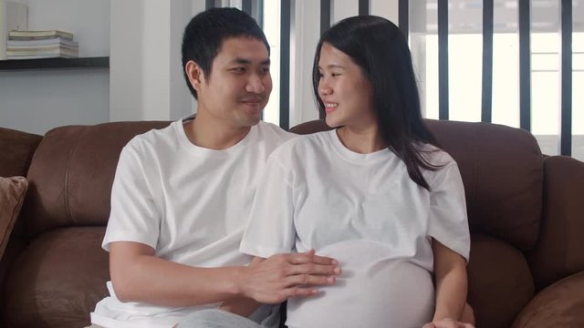 Young Asian Pregnant couple making heart sign holding belly. Mom and Dad feeling happy smiling peaceful while take care baby, pregnancy lying on sofa in living room at home concept. Slow motion.