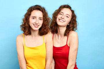 smiling twin sisters posing to the camera isolated on blue, beauty concept, girl in red T-shirt looks like the other one in yellow T-shirt.