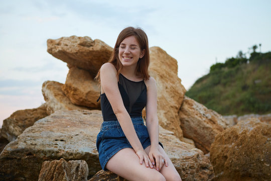 Young cheerful ginger cute lady with freckles sits on the beach, sitting on a stone, dreamily looks away and smiles, looks positive and happy.