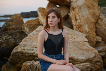 Photo of young redhead cute lady with freckles sits on the beach, dreamily looks away and smile, looks positive and happy.