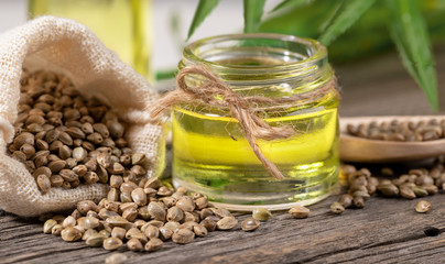 CBD oil.  Close-up glass jar with hemp oil and cannabis seeds in sack and in wooden spoon on old wooden board.