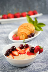 white bowl of porridge with strawberries and cherries, croissants and crispbread with cheese and strawberries on a gray background