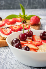 white bowl of porridge with strawberries and cherries, croissants and crispbread with cheese and strawberries on a gray background