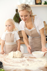 Little girl and her blonde mom in beige aprons playing and laughing while kneading the dough in kitchen. Homemade pastry for bread, pizza or bake cookies. Family fun and cooking concept