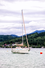 Fototapeta premium A white yacht in calm Kammersee's water. A cloudy day at Attersee lake at Salzkammergut area in Austria. There are a lot of green trees in the Alps mountains. The sky is full of grey clouds.