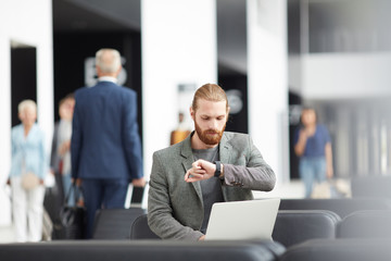 Serious young bearded entrepreneur sitting with laptop in airport and looking at wristwatch while...