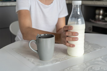 There is a gray cup with freshly prepared morning coffee on a white table in the kitchen . A woman puts a bottle of milk on the table after preparation. 