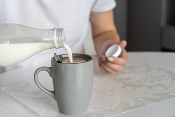 Close-up. On a white table in the kitchen there is a gray cup with freshly made morning coffee. A woman pours milk from a bottle into a cup.