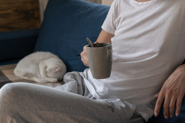 A woman is sitting at home on a sofa and holding a mug of coffee in her hand. Little white cute cat sleeping on the sofa in the background. Comfortable rest at home. 