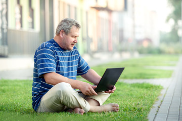 Beautiful adult man sitting on the lawn in the city with laptop. Of modern urban life.