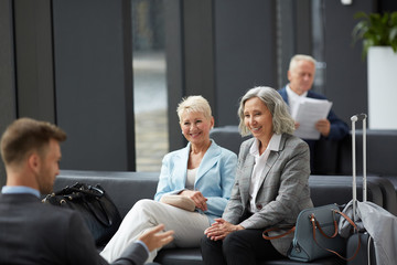 Cheerful mature business colleagues in jackets sitting on sofa in waiting area of airport and...