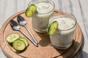 On a table on a wooden stand are two glass cups with a smoothie with cucumber and greens and metal spoons lie. Close-up.