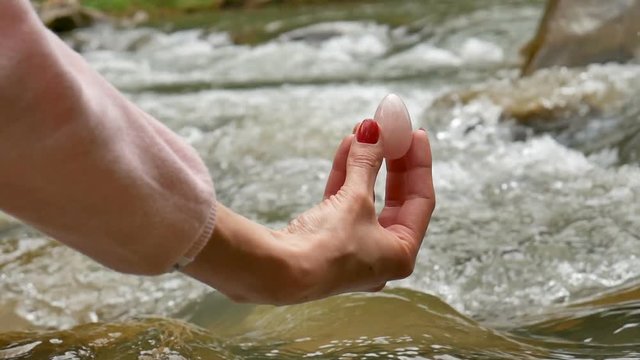 Female hand holding a rose quartz crystal yoni egg on river background. Women's health, unity with nature concepts.