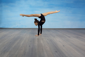 Fototapeta premium Girl gymnast in a black gymnastic swimsuit makes handstand legs divorced in splits on a blue sky background.