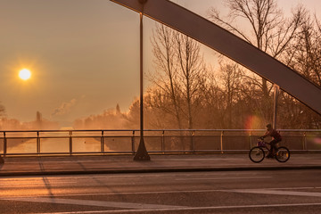 View through a bridge bow to a sunrise over a channel in Berlin on a misty morning with fairytale sunlight. On the right side you can see a cyclist.