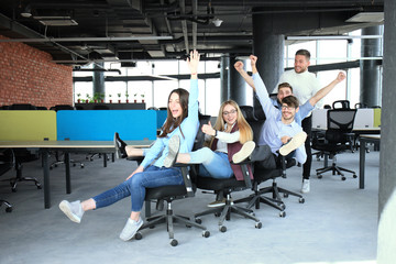 Young cheerful business people in smart casual wear having fun while racing on office chairs and smiling.