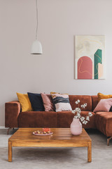 Flowers in vase on wooden coffee table in fashionable living room interior with brown corner sofa with pillows and abstract painting on the wall