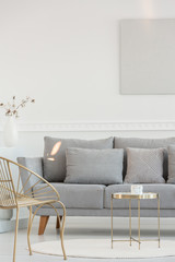 Stylish golden armchair and coffee table in front of grey comfortable sofa with pillows, copy space on empty white wall