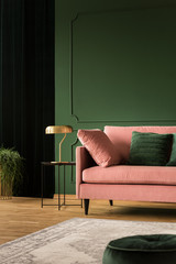 Stylish golden lamp on industrial table next to pink couch with emerald pillows in dark green...