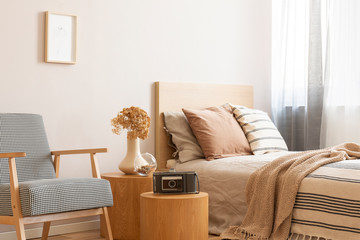 Vintage camera on round wooden table in the middle of elegant bedroom with retro armchair and single bed with beige bedding and flowers in vase