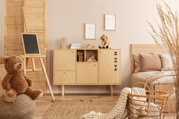 Teddy bear on pouf and small blackboard on easel in stylish kid's bedroom with wooden cabinet and...