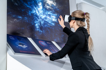 A girl in a black jacket standing in front of control panel monitors in virtual reality glasses. Looking forward, holds hands in front of the panels. Middle shot. Spaceship interior. Low angle