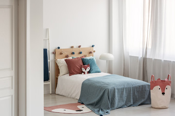 Bright bedroom interior with single bed with turquoise blanket on white bedding and colorful pillows and toy