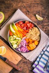 Bowl of salad with quinoa, carrots, avocado, onion, pepper, tomato with lime slices on a linen napkin on a wooden rustic table, top view