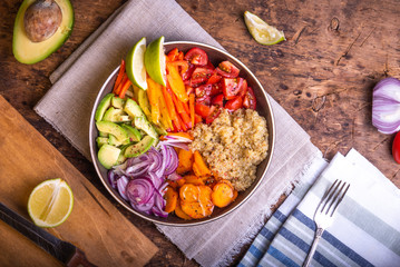 Bowl of salad with quinoa, carrots, avocado, onion, pepper, tomato with lime slices on a linen napkin on a wooden rustic table, top view