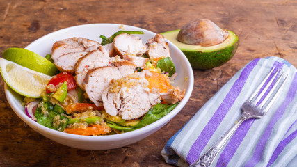 Bowl of salad with quinoa, carrots, avocado, onion, pepper, tomato with lime slices and chopped chicken breast on a rustic wooden background with ingredients - healthy eating concept