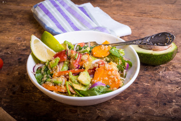 Bowl of salad with quinoa, carrots, avocado, onion, pepper, tomato with lime slices on a rustic wooden background