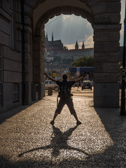 Fun in Prague - silhouette of a man posing beneath an arcade. He is turning his back to camera and throwing his shadow on a sidewalk in a stark back light. Sun lit Prague castle in distance.