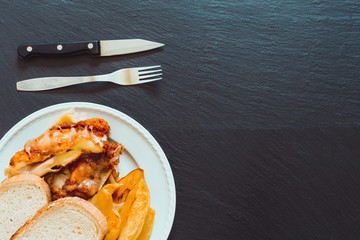 Fried chicken meat with poached potatoes next to cutlery with copy space – Food background made of meal next to cutlery on a dark black slate of ardesia