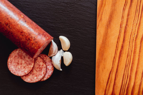 Salami With Slices And Garlic Cloves Next To An Empty Wooden Cutting Board – Traditional Gourmet With Chopped Pork Meat Inside Casing