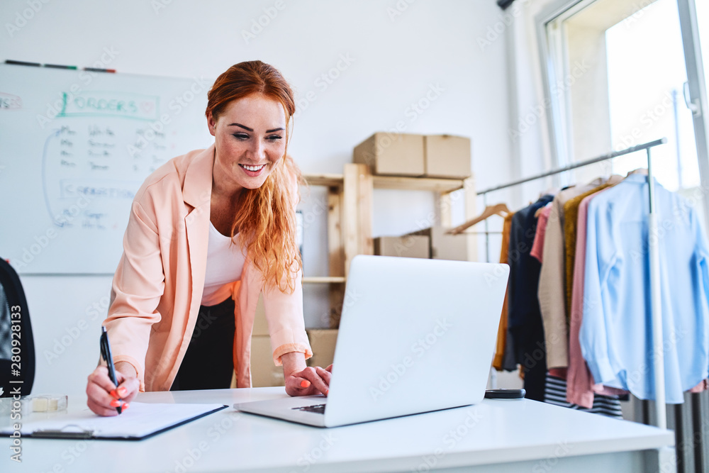 Wall mural cheerful female business owner looking at laptop and marking sales and deliveries on clipboard