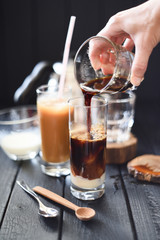 Making iced coffee in tall glasses. Woman hand pouring black coffee over ice cubes and condensed milk on black background