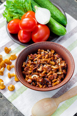 Fried chanterelles with onion in rustic bowl and plate with fresh vegetables for salad on background