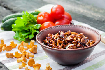 Fried chanterelles with onion in rustic bowl and plate with fresh vegetables for salad on background