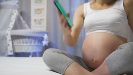 Future mother sitting on the bed and looking for information in her smartphone and shows a green screen in close-up, cellphone with chroma key and tracking motion markers. Baby crib on the background - Powered by Adobe