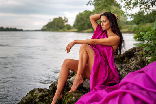 Artistically abstract image of a nude woman covered by a violet cloth. The sexy girl with long dark hair sits on the beach, in the background the water of the Danube river